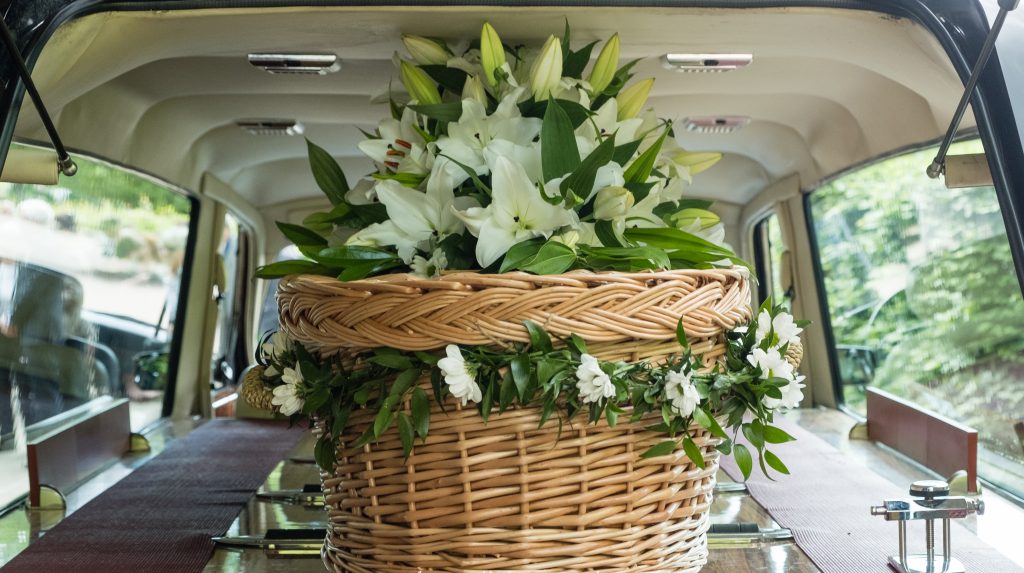 Wicker casket adorned with floral arrangement in back of hearse